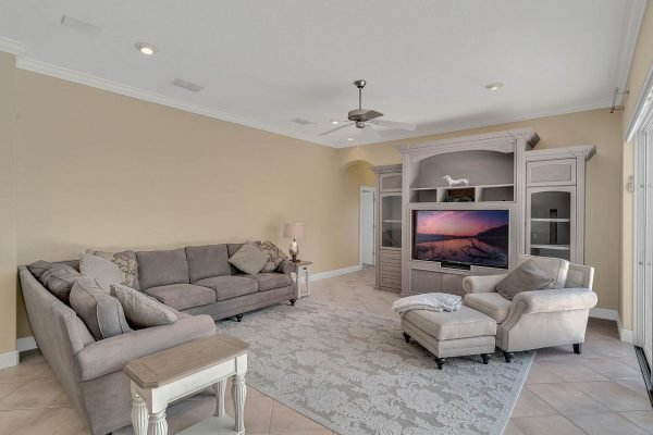 Spacious living room in a lakefront home in Windermere, featuring a gray sectional sofa, decorative pillows, a light-colored armchair, and a large entertainment center with a TV, all set against neutral walls and tiled flooring.