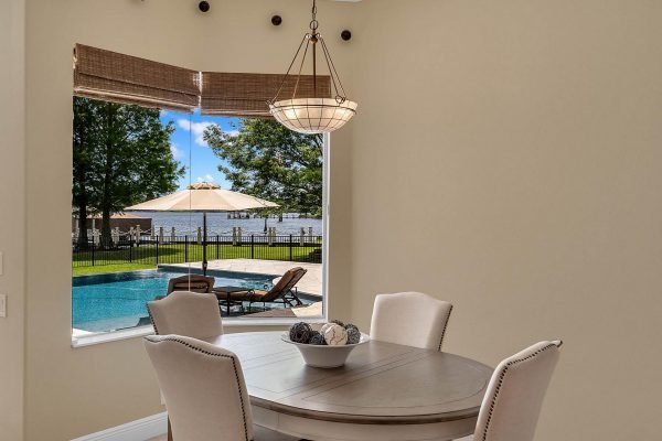 Dining area with a round table and chairs, large window showcasing a view of a private lake, outdoor pool, and patio area with lounge chairs and umbrellas, in a lakefront home in Windermere, Florida.