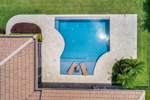 Aerial view of a private lakefront home in Windermere, FL, showcasing a saltwater infinity pool with a travertine patio, surrounded by lush greenery and landscaped gardens.
