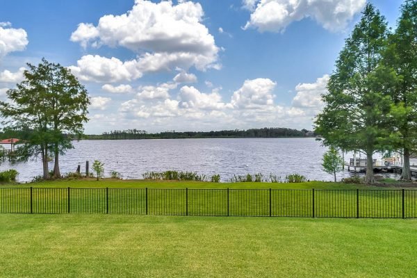 Lakefront view from a backyard in Windermere, Florida, featuring a fenced grassy area, trees, and a serene lake with a natural shoreline under a partly cloudy sky.