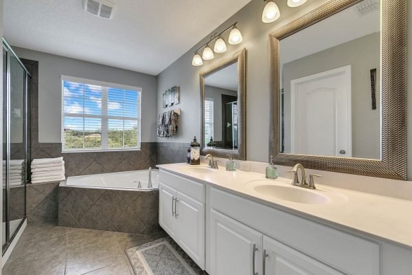 Master bathroom featuring dual sinks, large mirrors, a soaking tub, and a glass shower; bright natural light from window, elegant decor, and modern finishes.