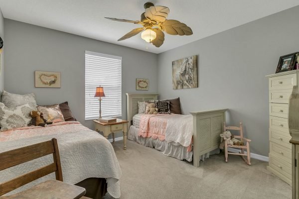 Cozy bedroom with two beds, decorative pillows, a bedside lamp, and a ceiling fan, featuring soft gray walls and natural light from a window, ideal for a family home at 2989 Park Meadow Drive, Apopka, FL.