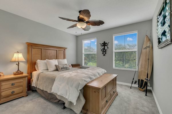 Cozy bedroom featuring a wooden bed with decorative pillows, bedside lamp, and a surfboard, complemented by natural light from two windows and a stylish ceiling fan.