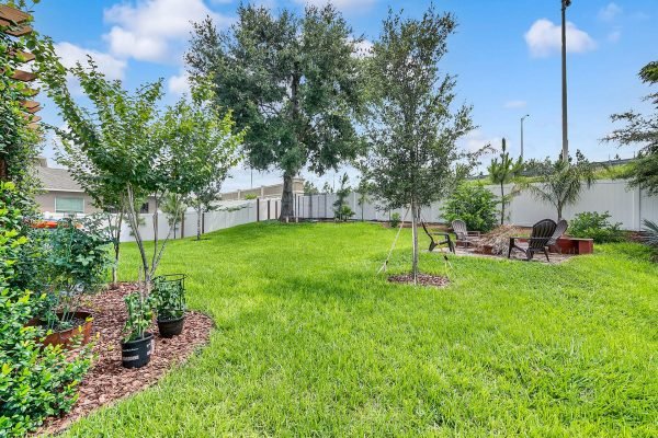 Backyard of 2989 Park Meadow Drive, Apopka, FL, featuring lush green lawn, mature oak tree, paver patio with fire pit, and seating area, surrounded by privacy fence and landscaped with small trees and plants.