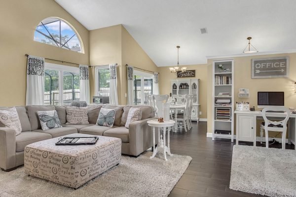 Spacious family room with a sectional sofa, decorative pillows, and a light-colored area rug, featuring large windows for natural light, a dining area, and a home office setup, reflecting a cozy and inviting atmosphere in a Lake Mary, FL home.