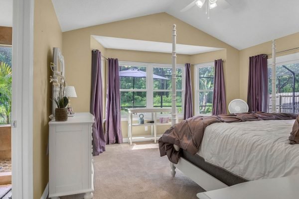 Master bedroom in Lake Mary, FL with vaulted ceiling, large windows, and purple curtains, featuring a cozy bed and white furniture, overlooking a serene backyard with conservation area.