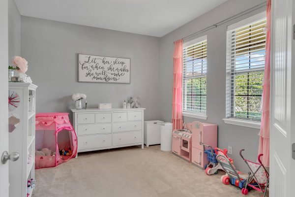 Child's bedroom with light gray walls, white furniture, pink curtains, and a play area featuring a toy kitchen and stroller, enhancing family-friendly appeal in Windermere home.