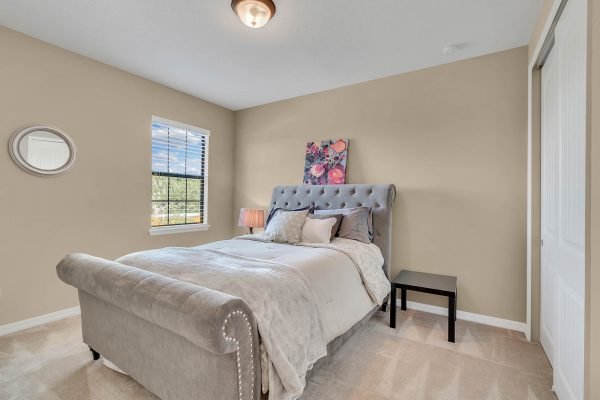 Cozy bedroom in Windermere home featuring a plush gray tufted bed, decorative pillows, and a bedside table with a lamp, complemented by a window allowing natural light and a floral artwork on the wall.