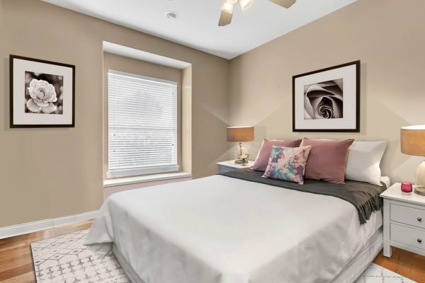 Cozy bedroom featuring a queen-sized bed with decorative pillows, framed floral artwork on beige walls, bedside tables with lamps, and a window with blinds, emphasizing a warm and inviting atmosphere.