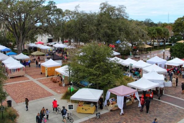 Winter Garden Farmers Market featuring numerous vendor tents, shoppers browsing, and lush greenery in a vibrant community setting.