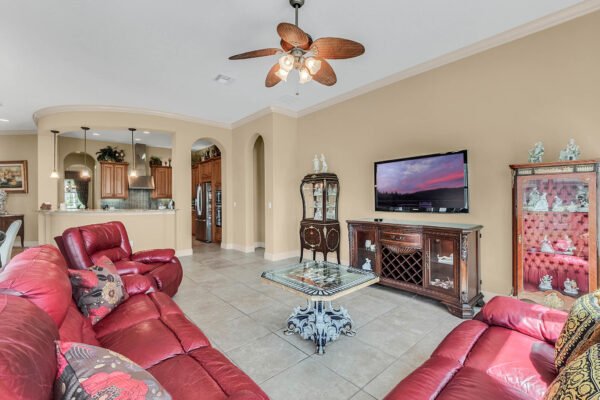 Living room featuring red leather sofas, a decorative coffee table, and a flat-screen TV, with an open layout connecting to the kitchen area, showcasing elegant furnishings and tile flooring, ideal for a luxury home listing at 1837 Lake Roberts Landing Dr, Winter Garden, FL.