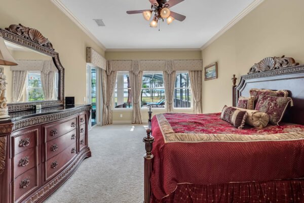 Master bedroom featuring a luxurious red bedspread, ornate wooden furniture, large windows with lake views, and elegant decor in a lakefront property at 1837 Lake Roberts Landing Dr, Winter Garden, FL.