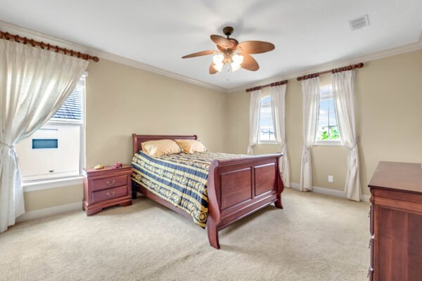 Spacious bedroom featuring a wooden bed with decorative bedding, two windows with sheer curtains, a ceiling fan, and a matching wooden dresser, showcasing the interior design of the luxury home at 1837 Lake Roberts Landing Dr, Winter Garden, FL.