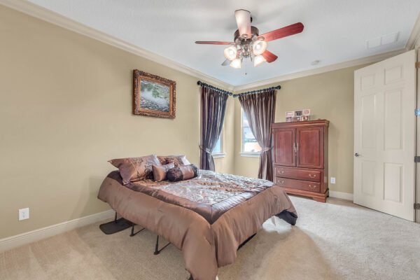 Cozy bedroom featuring a queen-sized bed with decorative pillows, wooden dresser, large windows with curtains, and ceiling fan, showcasing a warm and inviting atmosphere in the luxury home at 1837 Lake Roberts Landing Dr, Winter Garden, FL.