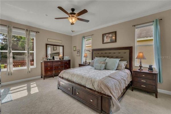 Spacious master bedroom featuring a king-size bed with decorative pillows, wooden furniture including a dresser and nightstands, large windows allowing natural light, and a ceiling fan, located in a home in Winter Garden, FL.