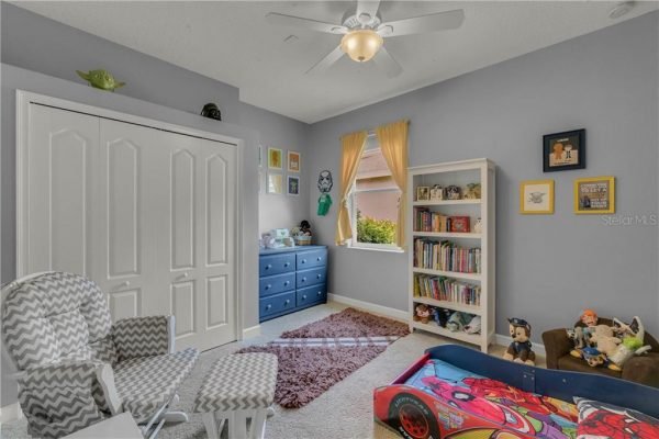 Child's bedroom featuring a blue dresser, bookshelf filled with toys and books, cozy seating area with patterned chairs, and bright window with yellow curtains, ideal for family living in Winter Garden, FL.