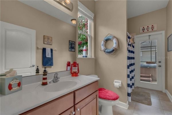 Bright and airy bathroom with beige walls, wooden cabinetry, decorative nautical elements, and a window overlooking a backyard, featuring a shower curtain with blue stripes and a life preserver.
