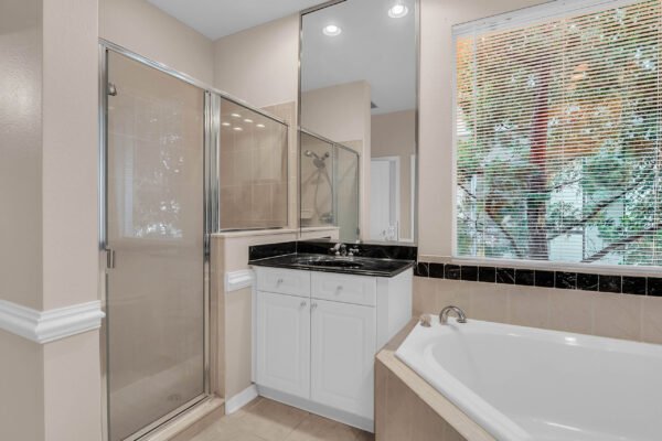 Bathroom with glass shower enclosure, white cabinetry, black countertop, and a large soaking tub, featuring natural light from window with blinds, reflecting a modern design suitable for a Windermere home listing.