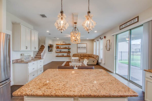 Modern kitchen and living area featuring an island with granite countertop, pendant lighting, white cabinetry, and a cozy seating area with decorative accents in a newly built home in Winter Garden, Florida.