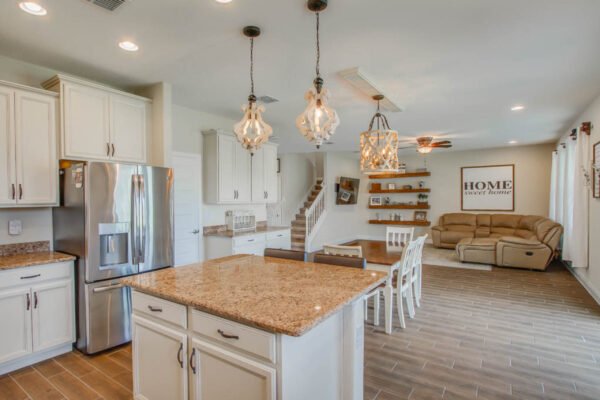 Modern kitchen with granite countertops, stainless steel appliances, and pendant lighting, featuring an open floor plan leading to a cozy living area with a "HOME sweet home" sign and comfortable seating.