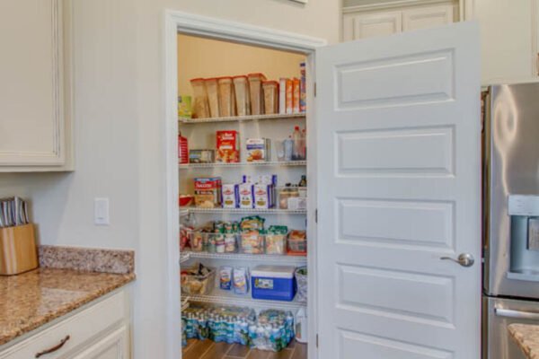 Pantry with organized shelves displaying various food items, including canned goods and snacks, adjacent to a kitchen with granite countertops and a stainless steel refrigerator.