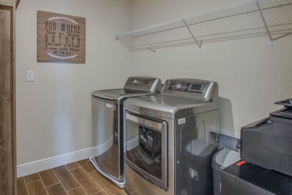 Laundry room with stainless steel washer and dryer, wall-mounted shelf, and decorative sign reading "For where two or three gather," emphasizing modern home amenities in Winter Garden, FL.