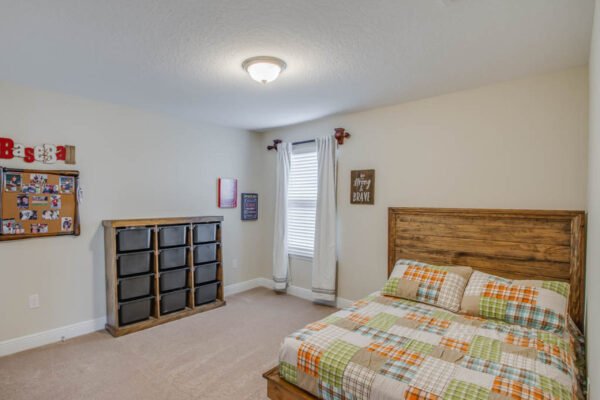 Cozy bedroom featuring a wooden bed with a colorful plaid comforter, a dresser with storage bins, a bulletin board, and natural light from a window in a modern home, ideal for families in the Waterleigh community near Winter Garden, FL.