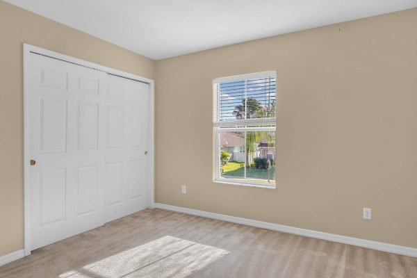 Bright bedroom with beige walls, carpeted floor, and window showcasing outdoor greenery, featuring double closet doors.