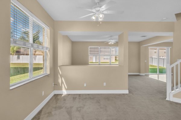 Interior view of a spacious living area in a Winter Garden home, featuring large windows, neutral walls, and carpet flooring, with a glimpse of the backyard and an open layout connecting to other rooms.
