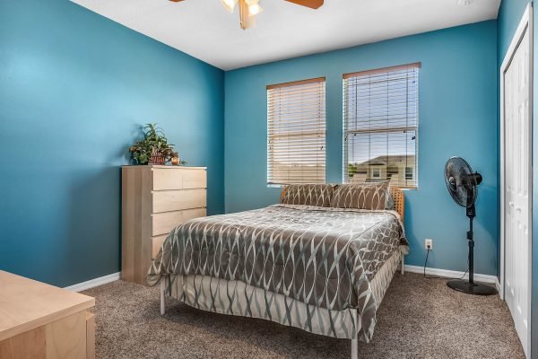 Cozy bedroom featuring a bed with a patterned comforter, a dresser, and a fan, set against a vibrant blue wall with natural light from two windows, ideal for showcasing the inviting interior of 11717 Sheltering Pine Drive, Orlando, FL.
