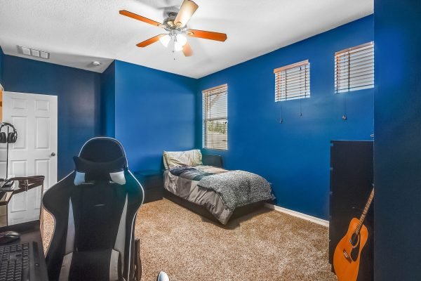 Bedroom with blue walls, a bed with gray bedding, a desk with a gaming chair, and a guitar against the wall, featuring windows with blinds and carpeted flooring.
