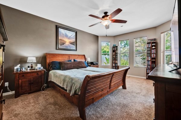 Spacious master bedroom featuring a wooden bed frame, plush carpet, and large windows with natural light, complemented by a ceiling fan and tasteful decor.
