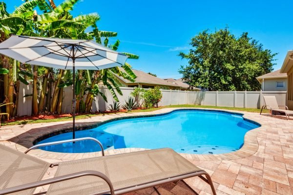 Backyard pool area featuring a saltwater pool, sun loungers, and an umbrella, surrounded by tropical plants and a fenced yard, ideal for relaxation in Florida.