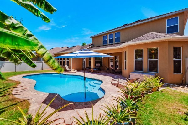 Backyard view of a spacious pool area with a blue umbrella, surrounded by lush greenery and tropical plants, featuring a well-maintained home in Orlando, Florida.
