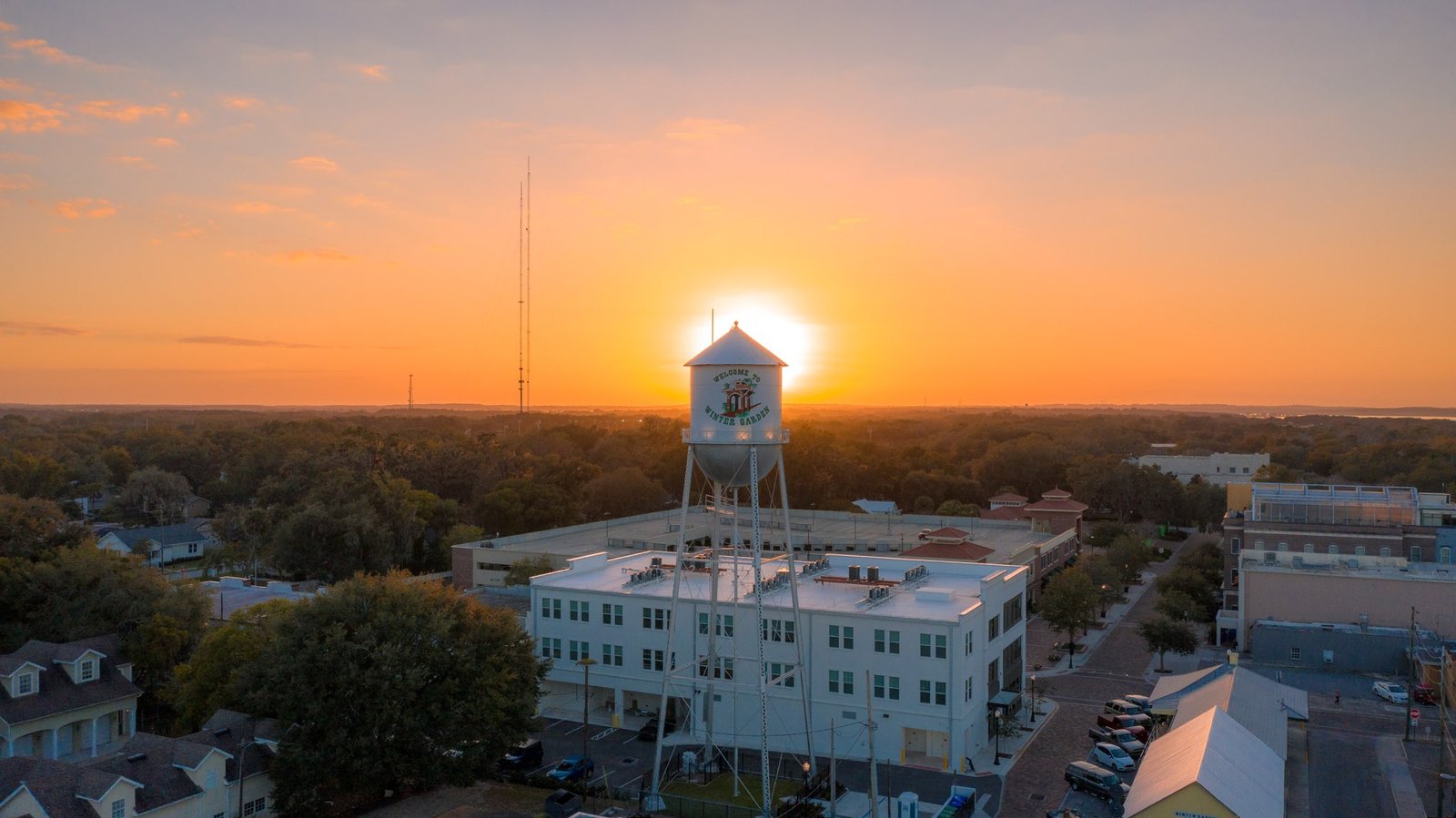 Water tower with "Welcome to Winter Garden" sign, illuminated by sunset, surrounded by trees and buildings in Winter Garden area.