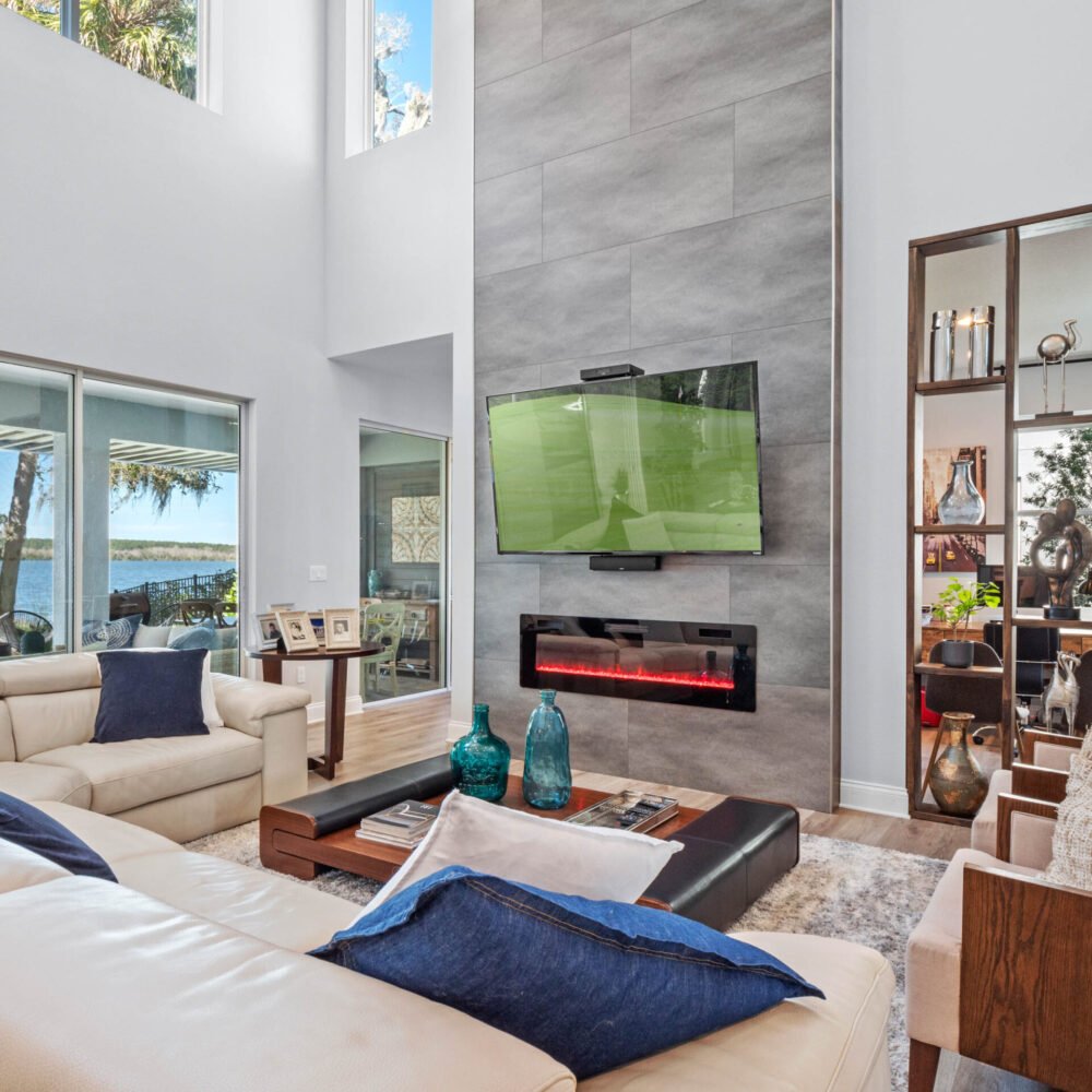 Modern living room with high ceilings, large windows showcasing lake views, sleek gray accent wall with mounted TV and electric fireplace, white leather sofas with blue and gray throw pillows, and stylish decor elements.