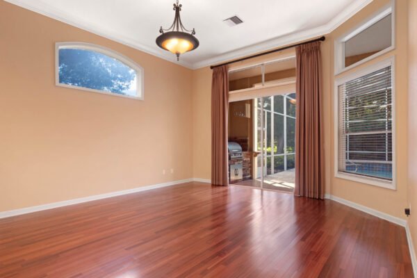 Spacious living room with hardwood flooring, large windows, and access to outdoor pool area, featuring elegant light fixture and warm beige walls, located in custom home at 11359 Willow Gardens Drive, Windermere.