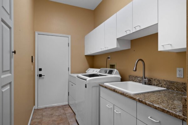 Laundry room with white cabinetry, granite countertop, washer and dryer, and a utility sink against a warm beige wall, featuring a door leading outside.