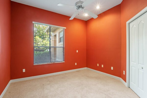Empty room with orange walls, ceiling fan, and carpet, featuring a window with blinds allowing natural light, and a door leading to another area.