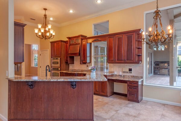 Elegant gourmet kitchen with dark wood cabinetry, granite countertops, stainless steel appliances, and a spacious island, featuring a chandelier and views of the outdoor living space.