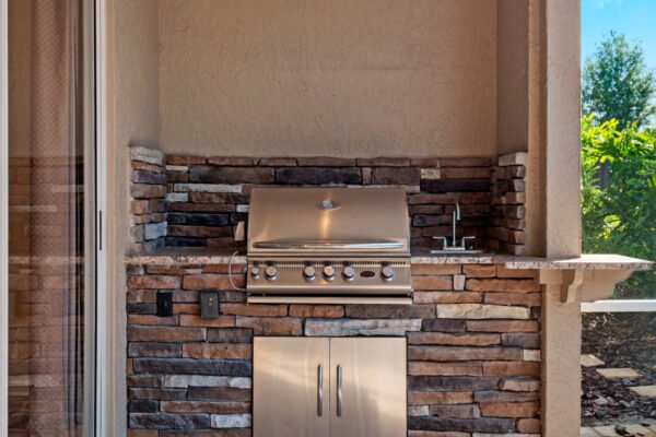 Outdoor summer kitchen with built-in stainless steel grill and stone countertop, featuring a sink and cabinetry, located in a Windermere home’s lanai area.
