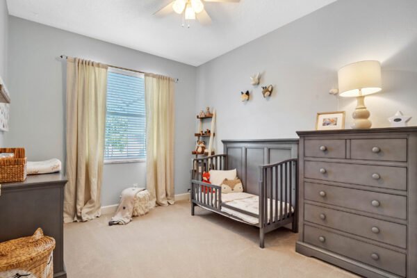 Cozy nursery featuring a gray crib, matching dresser, and soft beige carpet, illuminated by natural light from a window with sheer curtains, showcasing a warm and inviting atmosphere.