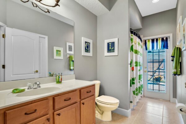 Bathroom featuring a light wood vanity with sink, decorative wall art, bright shower curtain with green and blue patterns, and a window with blinds showcasing outdoor views, highlighting the spacious and well-lit layout of the home at 13915 Zephermoor Lane in Winter Garden, FL.