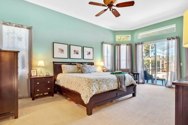 Spacious master bedroom with a king-sized bed, light green walls, and large windows showcasing pool views, featuring a wooden dresser, bedside lamps, and decorative wall art, ideal for a home in Winter Garden, FL.