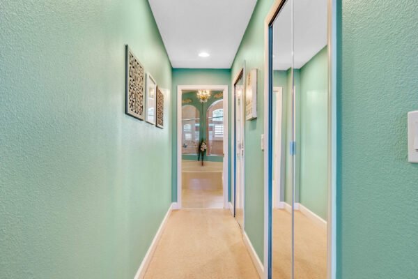 Hallway with teal walls, featuring decorative wall art, mirrored closet doors, and a view of a bright room with chandelier and plants, part of a 4-bedroom, 3-bathroom home in Winter Garden, FL.