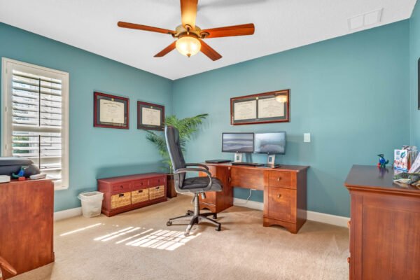 Home office with wooden desk and chair, blue walls, framed diplomas, two computer monitors, and natural light streaming through a window, showcasing a well-designed workspace in the Winter Garden property at 13915 Zephermoor Lane.