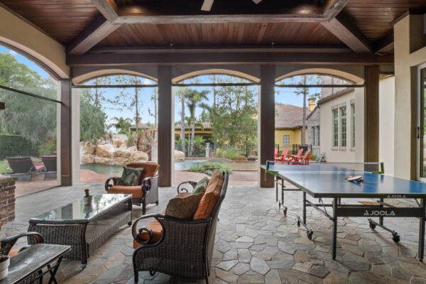 Luxury outdoor living space featuring a covered lanai with comfortable seating, stone flooring, and a ping pong table, overlooking a landscaped backyard with palm trees and a pool, ideal for enjoying the Florida lifestyle at 11019 Bridge House Road, Windermere.