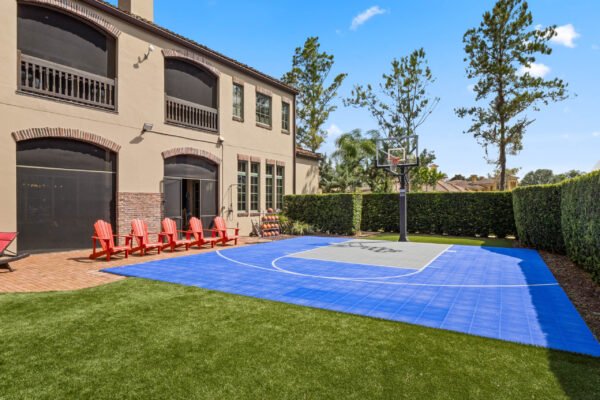 Luxury home backyard featuring a blue half-court basketball sport court, surrounded by manicured greenery, with red Adirondack chairs and a two-story residence in Windermere, FL.