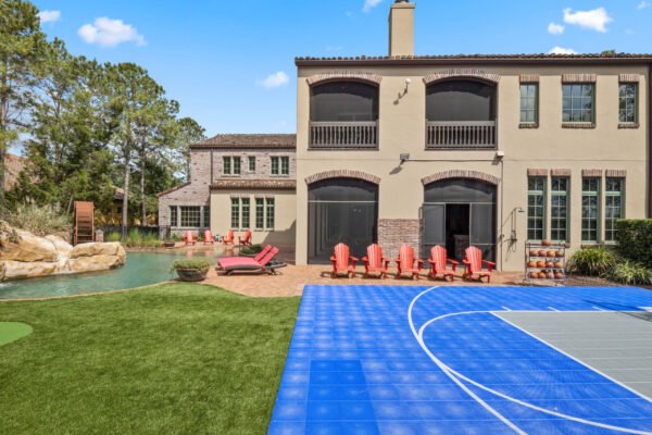 Lakefront luxury home at 11019 Bridge House Road in Windermere, featuring a spacious outdoor area with a blue basketball court, swimming pool, and lounge chairs, surrounded by lush greenery and stone accents.