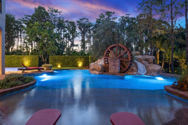 Luxury lakefront pool area featuring a water wheel, rock formations, and a waterslide, surrounded by lush greenery at 11019 Bridge House Road, Windermere, FL.